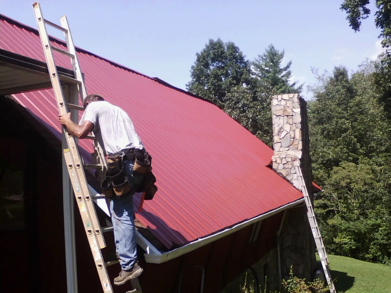 Roofer on a ladder beside a red metal roof and stone chimney — Soffit & Fascia Repair by Roofing Brothers in Clarington